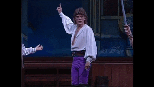 a man in a white shirt and purple pants is holding a sword on a stage and giving the middle finger .