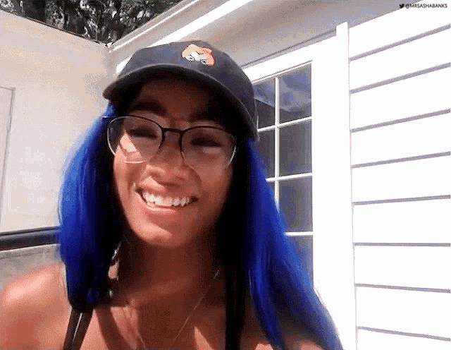 a woman with blue hair wearing glasses and a baseball cap smiles