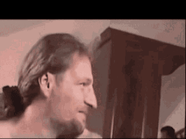 a man with long hair and a ponytail is standing in a room with a wooden cabinet in the background .