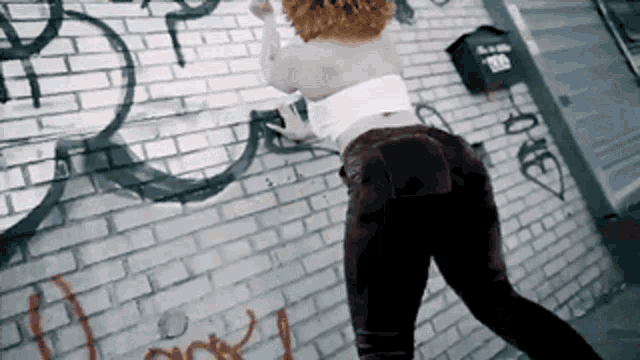 a woman is standing in front of a brick wall that has graffiti on it