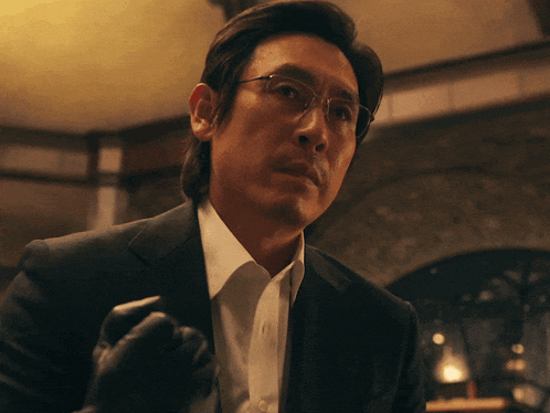 a man wearing glasses and a suit holds a glove in his hand