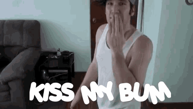 a man in a white tank top is standing in front of a couch and says kiss my bum
