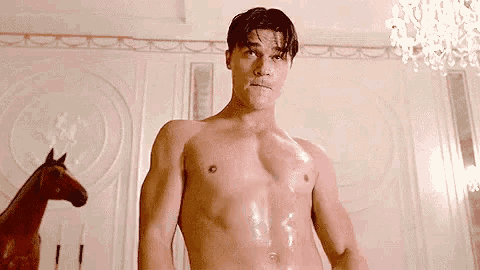 a shirtless man is standing in front of a pink wall in a room with a horse in the background .