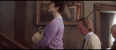 a woman in a purple sweater is standing next to a man and a woman in front of a painting