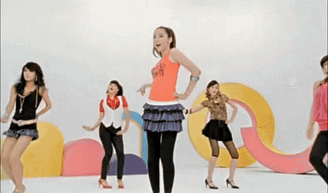 a group of young women are dancing in front of a colorful background and one of them is wearing an orange shirt that says i love ny