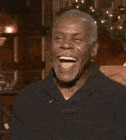 a man in a black shirt is laughing while sitting on a couch in front of a christmas tree