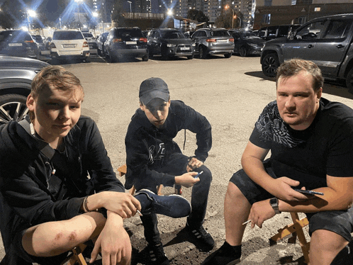 three men are sitting in a parking lot with one wearing a black sweatshirt with the letter f on the sleeve