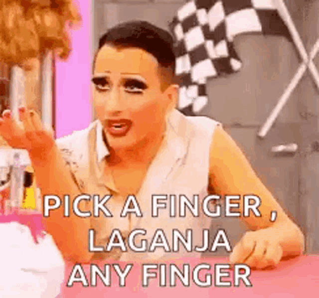 a drag queen is sitting at a table making a funny face and saying `` pick a finger , laganja any finger ''