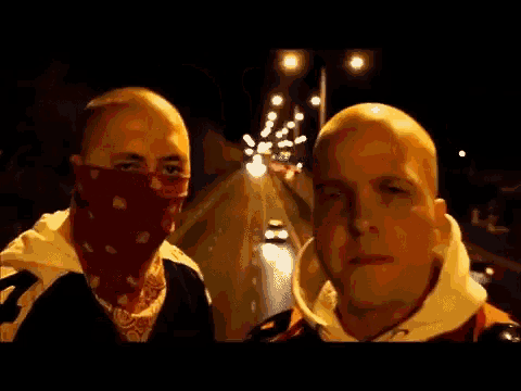 a man wearing a red bandana on his face stands next to another man
