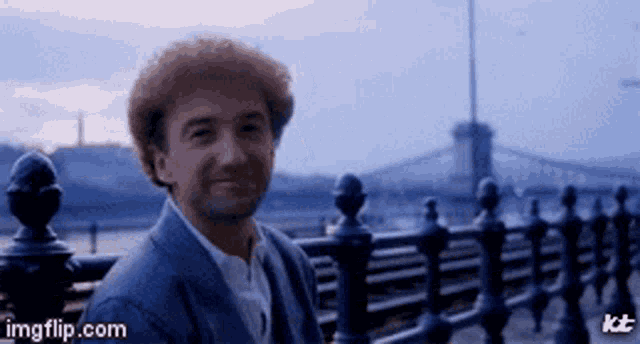 a man with red hair is smiling in front of a bridge and a fence .