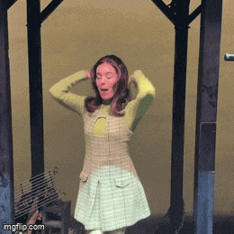 a woman in a plaid dress is standing in front of a wooden structure with her hands behind her head ..