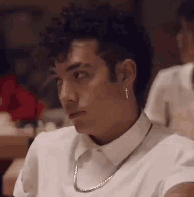 a man with curly hair and earrings is wearing a white shirt and a silver chain around his neck .