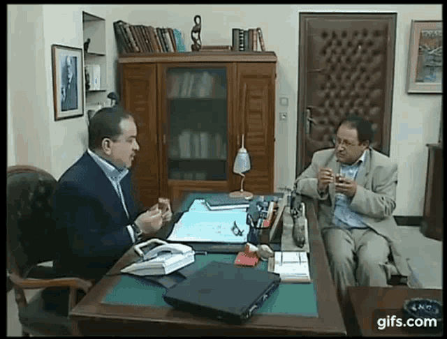 two men are sitting at a desk talking to each other in an office .