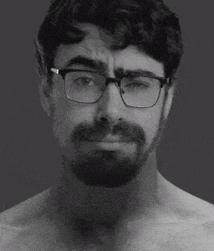 a shirtless man with glasses and a beard looks at the camera