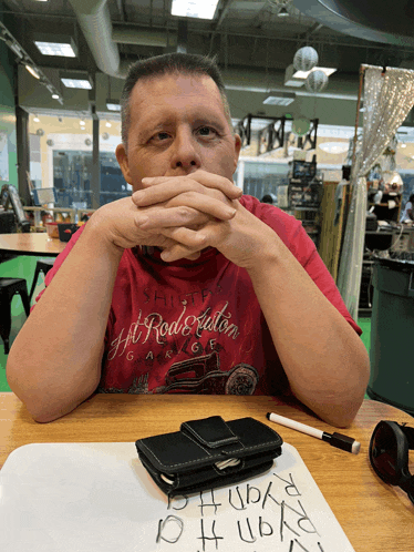 a man sits at a table with his hands on his face wearing a red shirt that says ' ship ' on the front