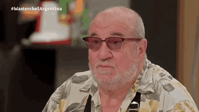 a bald man with a beard wearing sunglasses and a floral shirt is sitting in front of a sign that says masterchefargentina