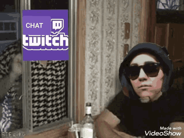 a man wearing sunglasses sits in front of a window with a twitch logo behind him