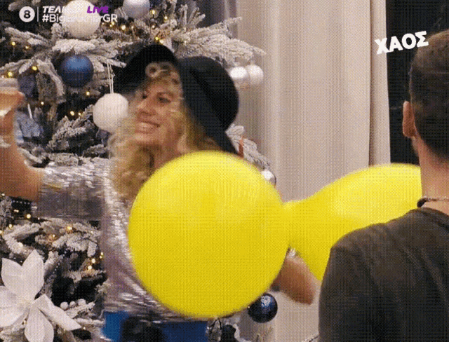 a woman in a black hat is holding two yellow balloons in front of a christmas tree ..