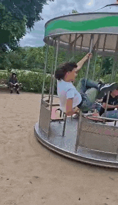 a man is riding a merry go round in a park while a woman sits on a bench in the background .