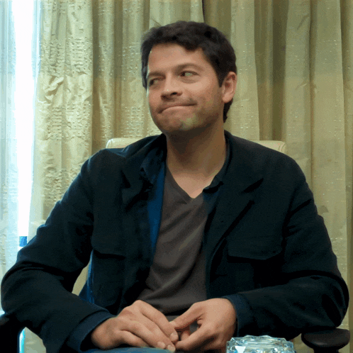 a man in a black jacket sits in a chair with his hands folded