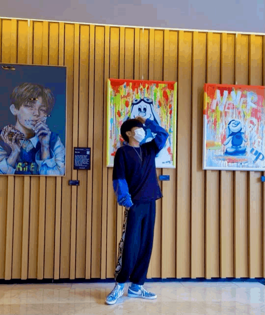 a man wearing a mask stands in front of a wall of paintings including one that says ' a ' on it