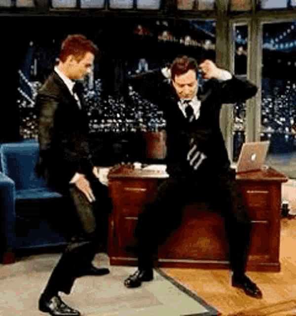 two men in suits are dancing in front of a desk with a laptop .
