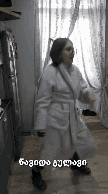 a woman in a white robe is dancing in a kitchen with a foreign language written on the bottom
