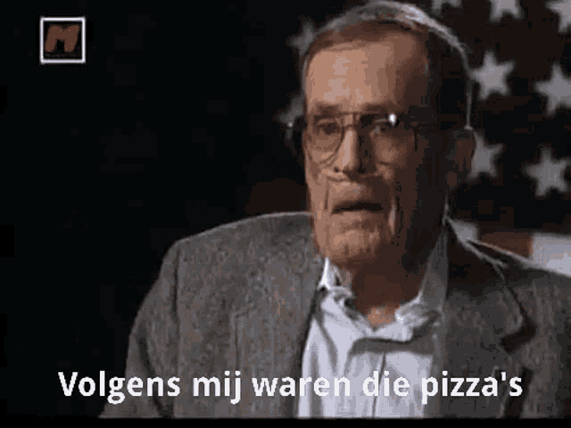 a man in a suit and glasses is standing in front of an american flag and says volgens mij waren die pizza 's .