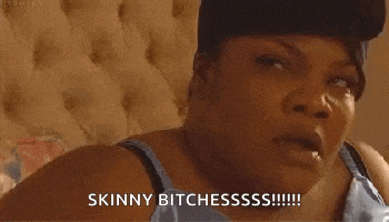 a woman wearing a hat and a blue tank top is sitting on a bed and says `` skinny bitchesssss ! ''
