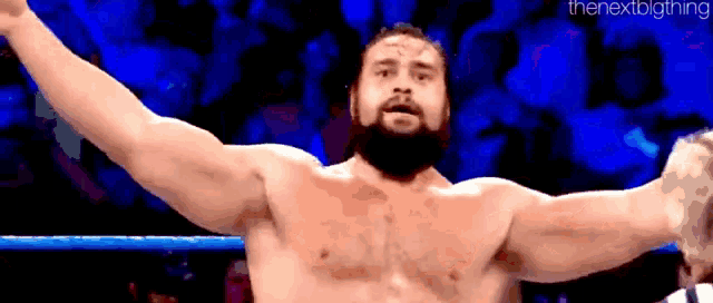 a shirtless wrestler with a beard is raising his arms in the air .