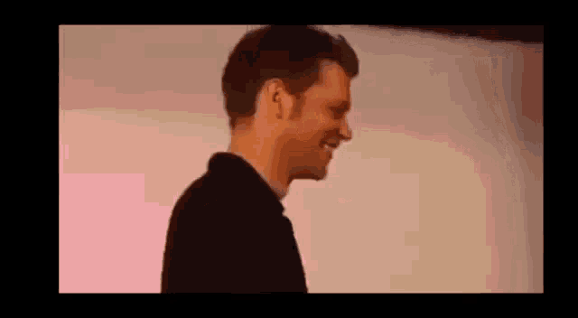 a man in a black shirt is smiling while standing in front of a pink background .