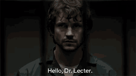 a man with curly hair and a beard is standing in a dark room and saying `` hello , dr. lecter . ''