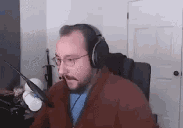 a man with a beard wearing headphones and glasses is sitting in front of a microphone in a room .