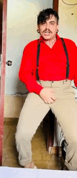 a man in a red shirt and suspenders is sitting on a chair with his hands on his hips .