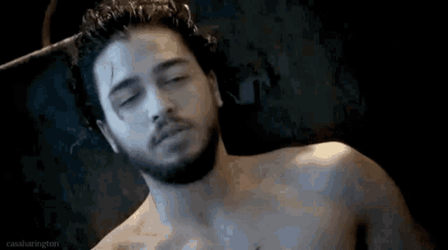 a shirtless man with a beard is laying down in a dark room with his eyes closed .