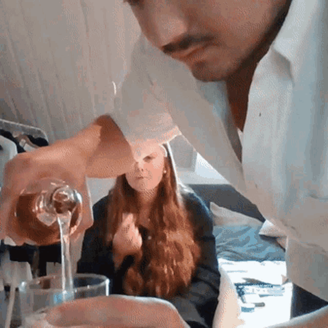 a man pours liquid into a glass while a woman sits behind him