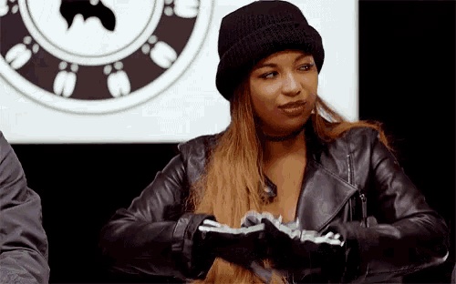 a woman wearing a black beanie and a leather jacket is sitting in front of a clock