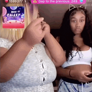 two women are standing next to each other in a room . one of the women is holding a cell phone .