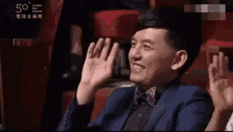 a man in a suit and bow tie is sitting in a theatre with his hands up .