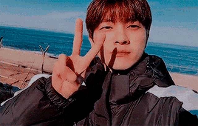 a young man in a black jacket is giving a peace sign on the beach .