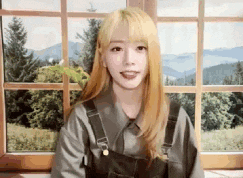 a young woman with blonde hair is standing in front of a window with trees in the background .