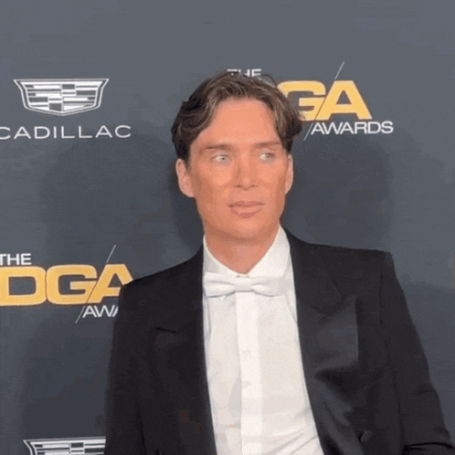 a man in a suit and bow tie is standing in front of a cadillac logo