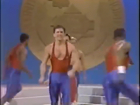a man in a red tank top and blue shorts is standing in front of a clock on a stage .