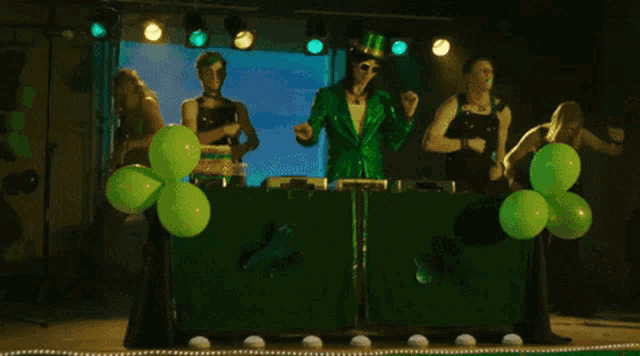 a group of people dressed in green are dancing in front of green balloons on a stage