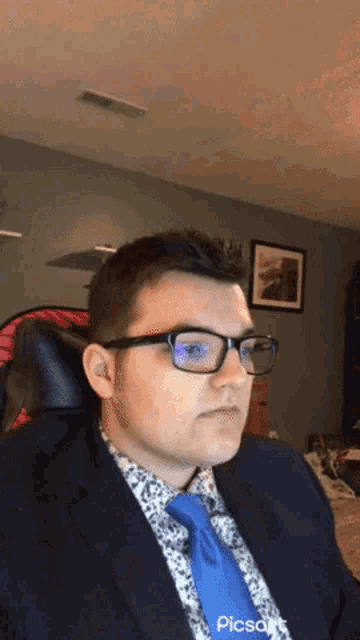 a man wearing glasses and a blue tie is sitting in a chair in a room