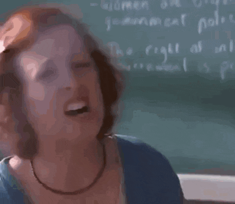 a woman with red hair is standing in front of a blackboard and making a funny face .