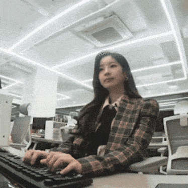 a woman in a plaid jacket sits at a desk typing on a keyboard