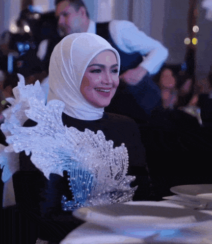a woman wearing a white hijab and a black dress smiles while sitting at a table