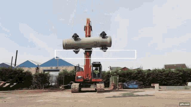 a large orange excavator is carrying a large pipe in a field