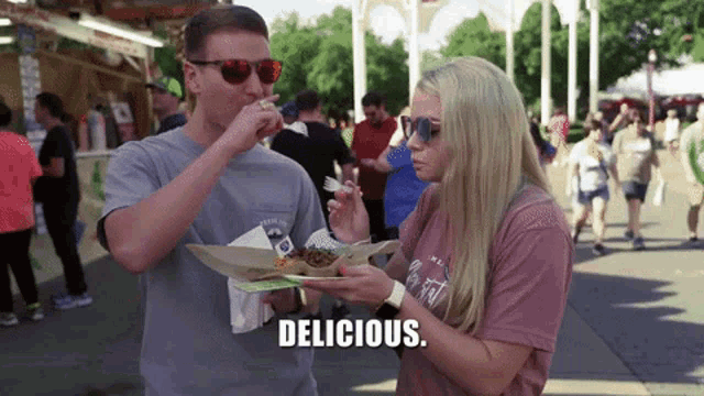 a man and a woman are eating food at a festival and the woman is saying delicious